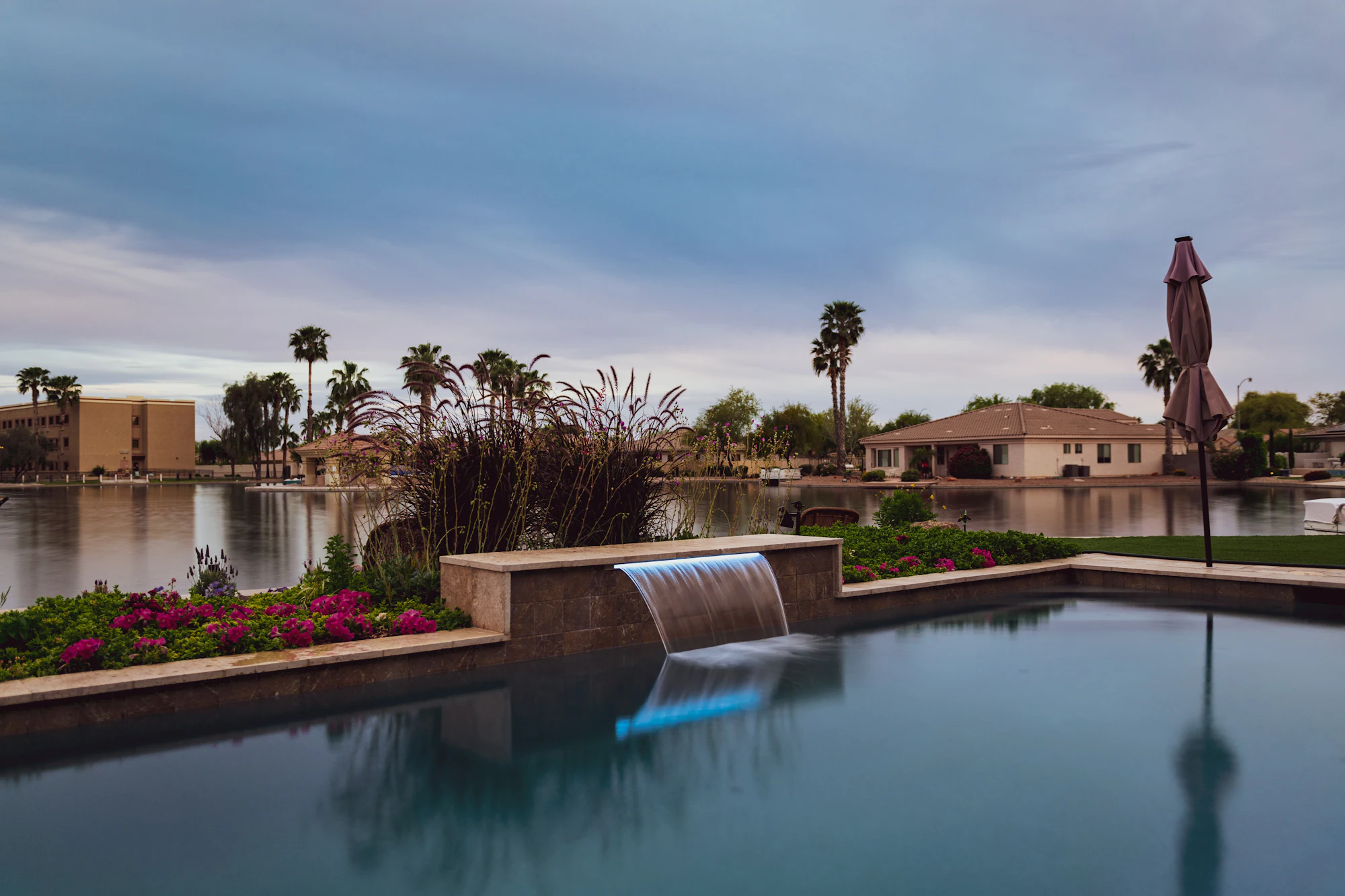 Resort-style community pool with a sheer waterfall, palms and homes across a lake at dusk